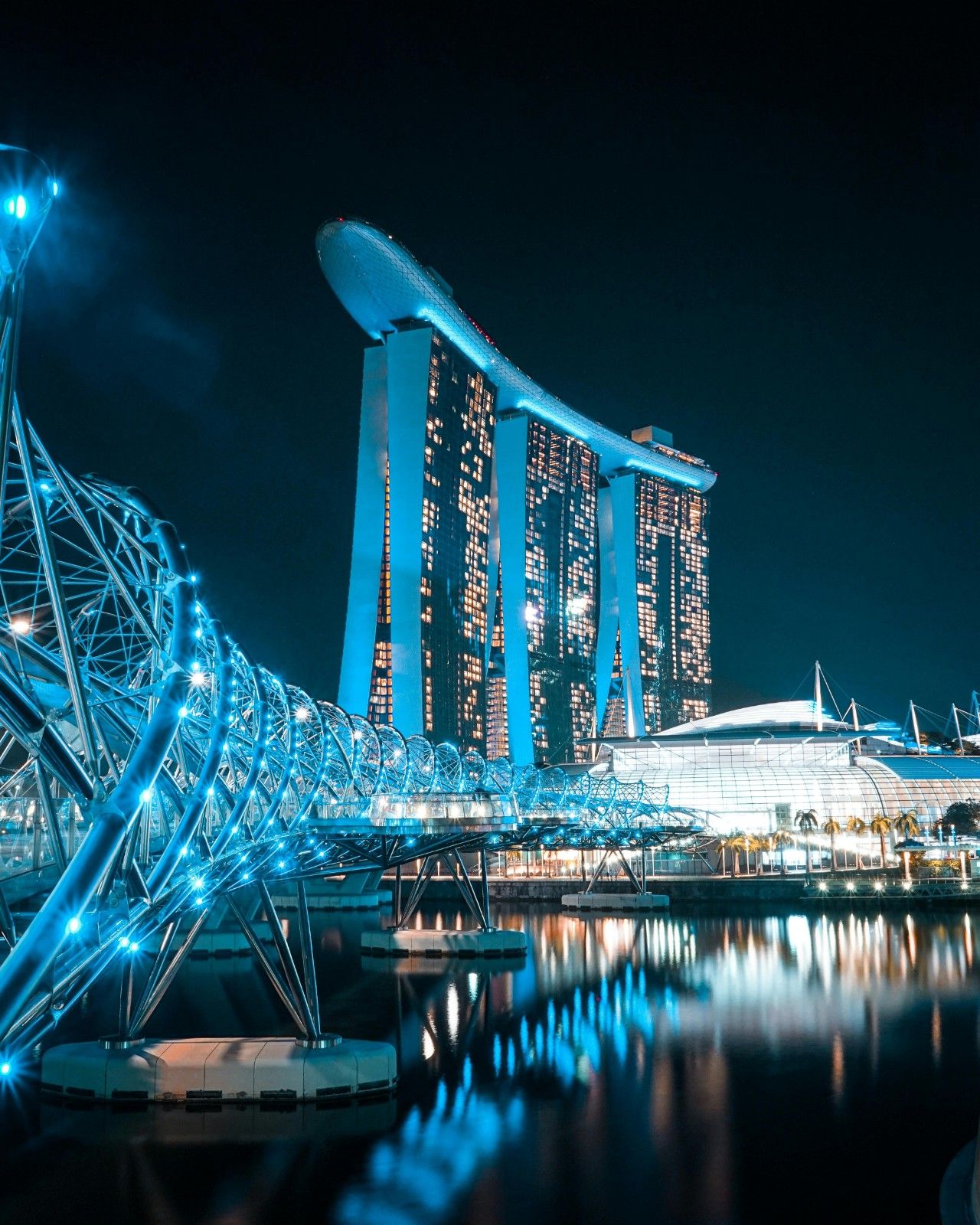 Nighttime view of the illuminated Helix Bridge and Marina Bay Sands hotel reflected in the water, Singapore.