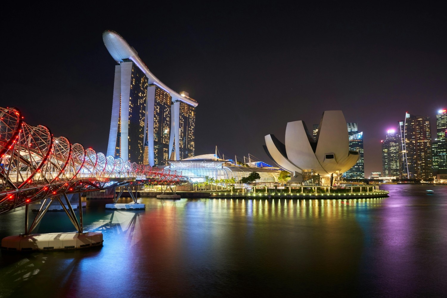 Nighttime view of the illuminated Helix Bridge and Marina Bay Sands hotel reflected in the water, Singapore.
