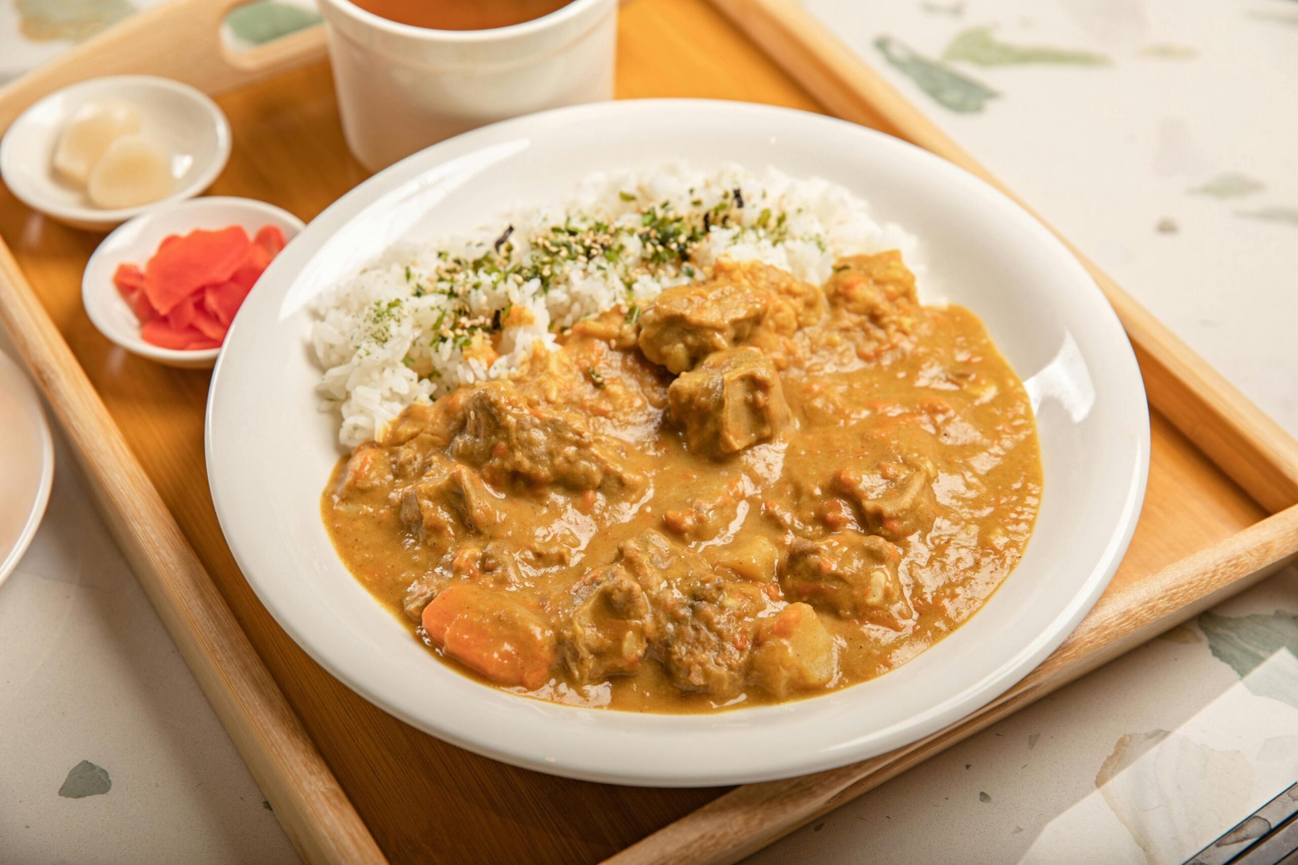 This image shows a hearty plate of Japanese-style curry with chunks of meat and carrots served alongside white rice topped with furikake seasoning. The meal is presented on a wooden tray with small side dishes of pickled ginger and radishes, accompanied by a cup of soup.