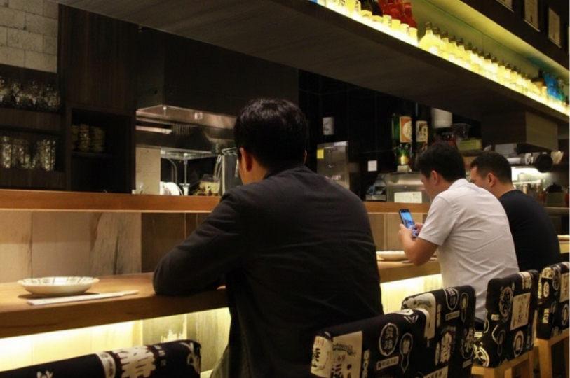 Three people sitting at a long wooden counter inside a modern Japanese izakaya, with glowing shelves of bottles and a kitchen area in the background.