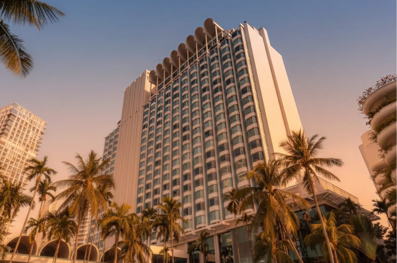 A low-angle shot of a tall, modern white hotel with distinct arched roof details, framed by palm trees against a soft sunset sky.