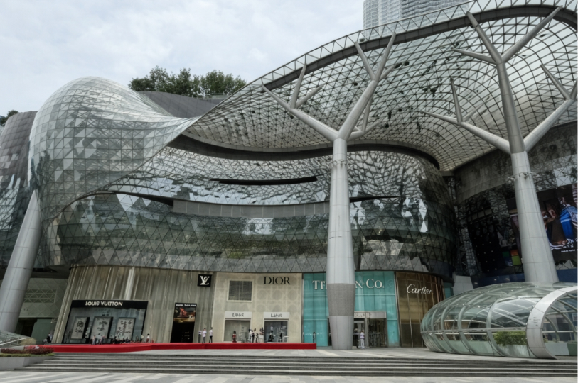 Modern, futuristic glass architecture of ION Orchard mall in Singapore featuring luxury storefronts like Louis Vuitton and Dior.
