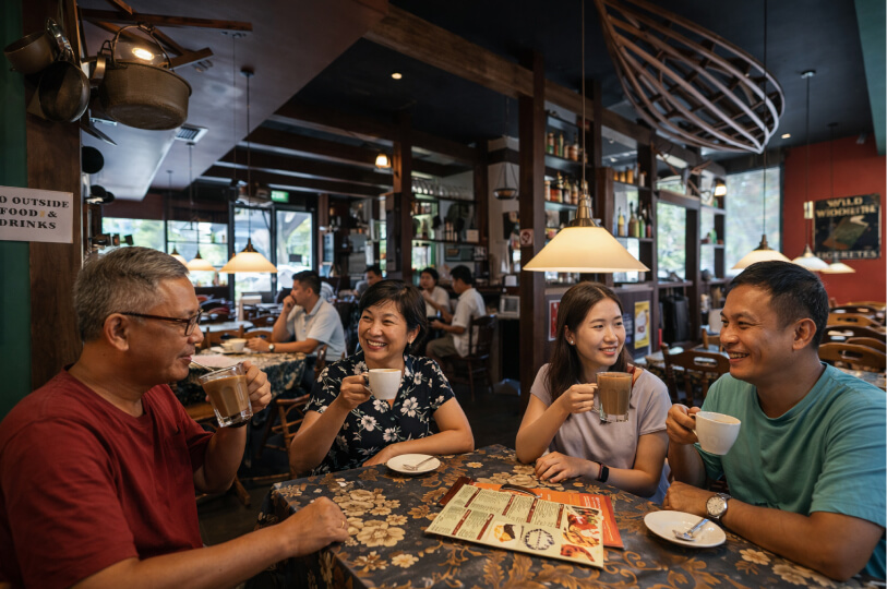 Four people sit at a floral-clothed cafe table, smiling and chatting over coffee. The interior features warm lighting, dark wood beams, and a decorative wooden boat frame hanging from the ceiling.