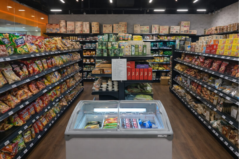 A wide-angle interior shot of a well-organized Korean grocery store. Two long aisles of snacks and dry goods frame a central freezer chest filled with frozen treats. The shelves are packed with colorful packaging, and the store is lit by modern linear ceiling lights.