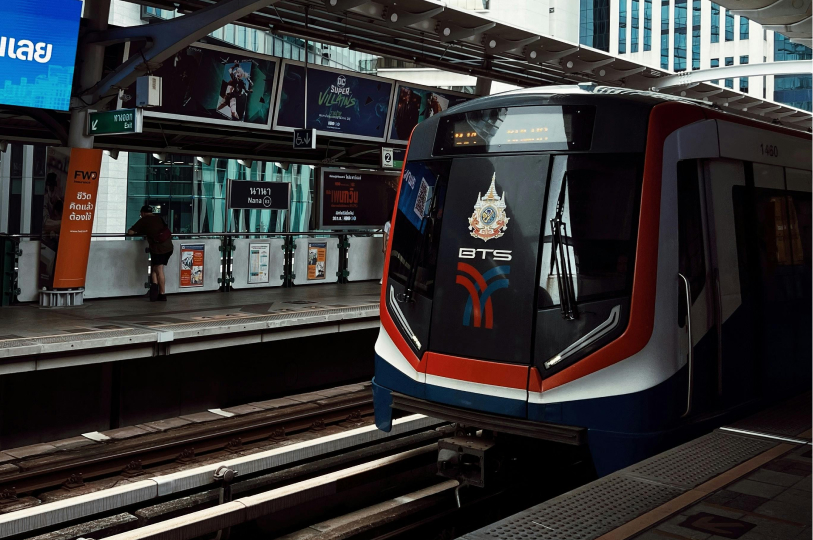A BTS Skytrain stopped at Nana Station in Bangkok, featuring the train's front with red, white, and blue branding alongside a person waiting on the platform.