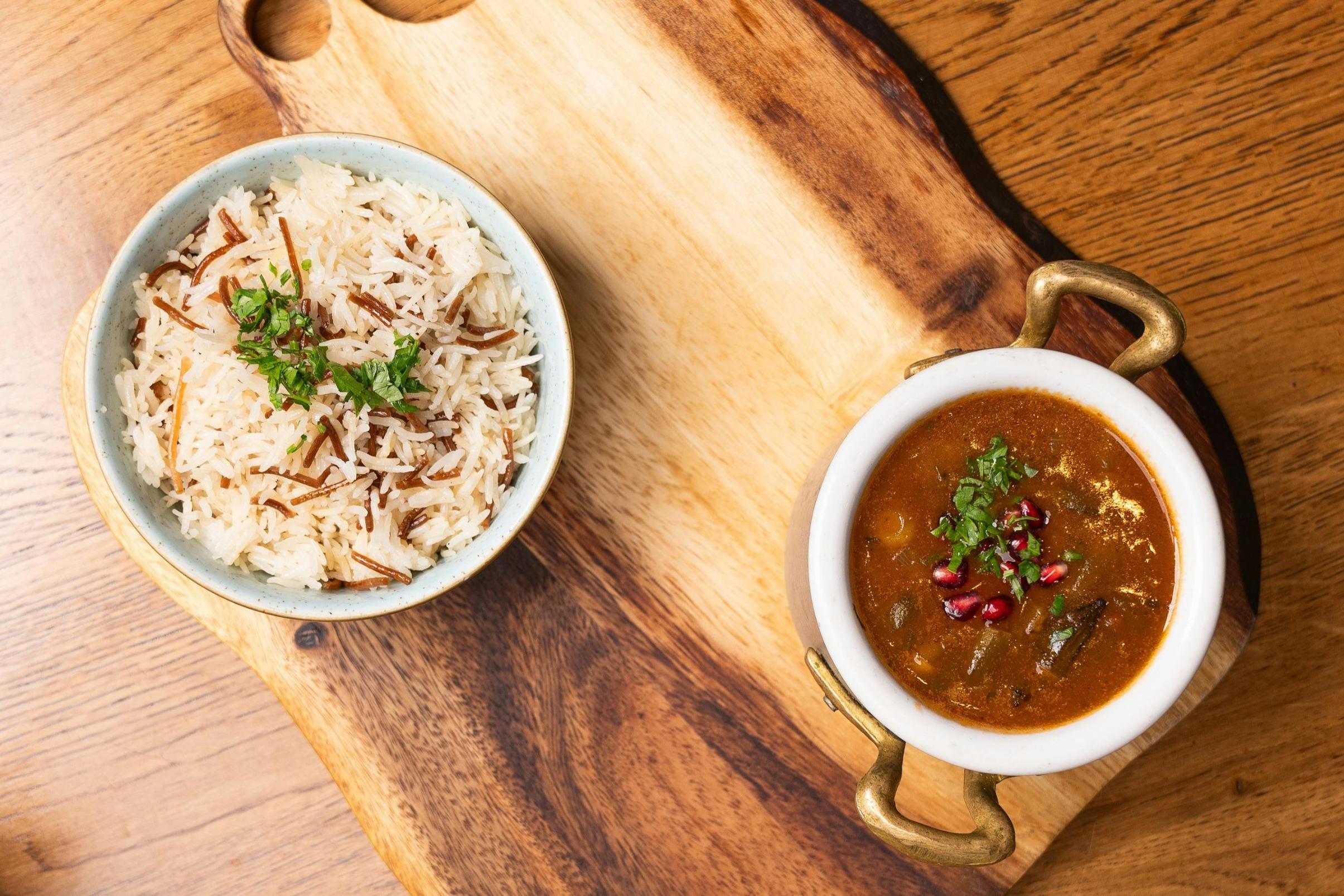  This image features a top-down view of a light blue bowl filled with rice pilaf mixed with vermicelli and fresh herbs sitting on a wooden cutting board. Next to it, a small white pot with brass handles contains a dark stew garnished with herbs and vibrant pomegranate seeds.