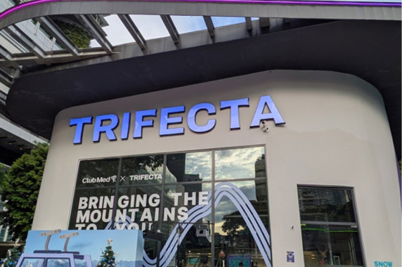 The exterior entrance of TRIFECTA Singapore with a large blue sign and a window display reading "Bringing the mountains to you."