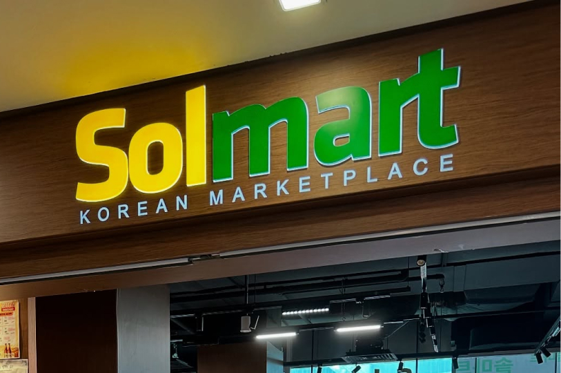 A close-up of a store sign on a wood-paneled wall that reads "Solmart KOREAN MARKETPLACE" in bold yellow and green 3D lettering. The lighting from above highlights the clean, modern design of the storefront.