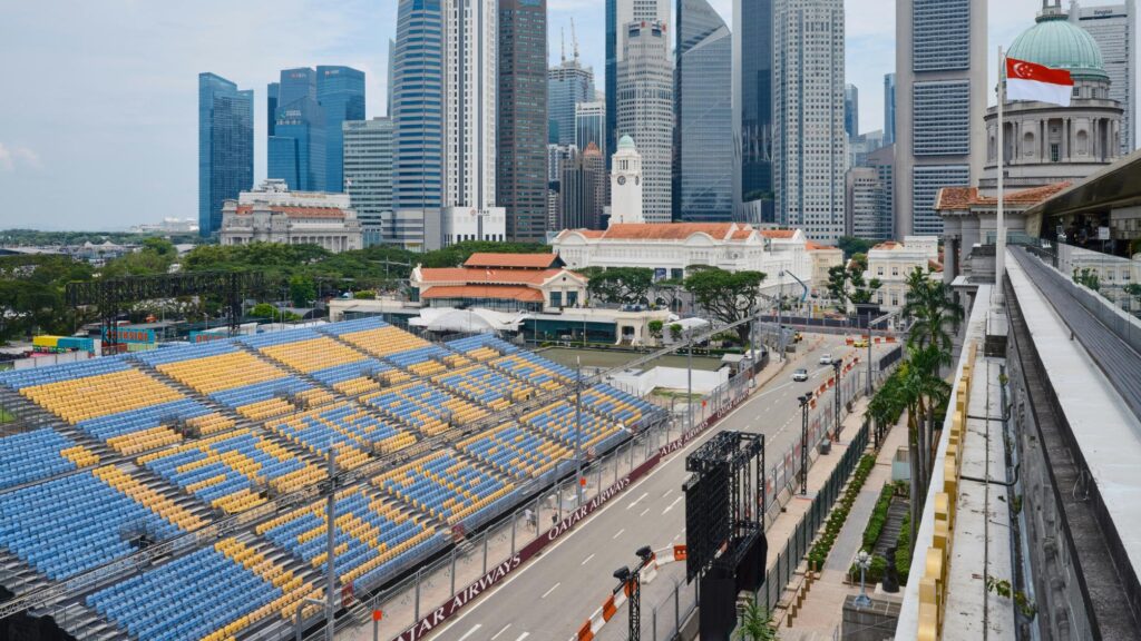 Rows of blue and yellow grandstand seating line a city street circuit bordered by fencing with "Qatar Airways" branding, indicating preparations for a major racing event. The scene captures a contrast between temporary sports infrastructure and the urban landscape, featuring historic colonial buildings and modern skyscrapers under a bright sky, with a Singapore flag flying in the foreground.