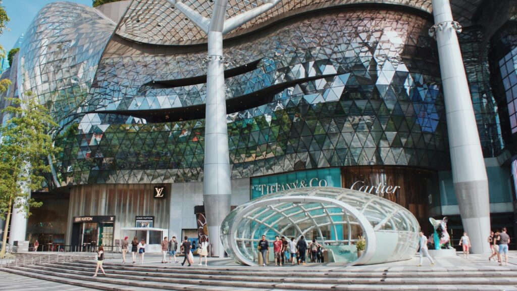 This modern architectural marvel features a sweeping, geometric glass façade supported by massive white pillars, housing high-end retailers like Louis Vuitton, Tiffany & Co., and Cartier. In the foreground, pedestrians stroll across a stepped plaza surrounding a distinctive, oval-shaped glass entrance structure.