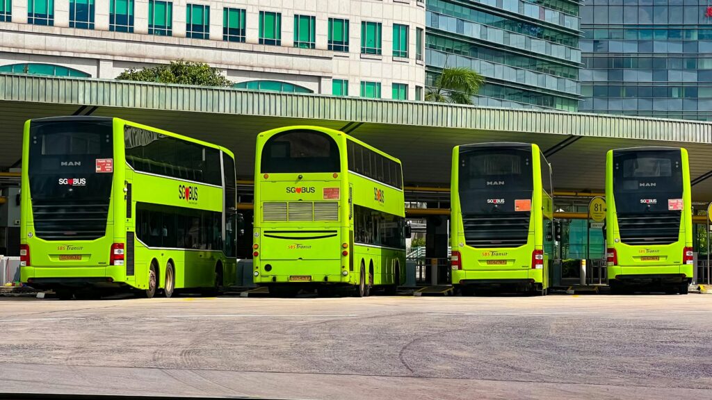 Four bright lime green double-decker buses, displaying "SG Bus" and "SBS Transit" branding, are parked in an orderly row beneath a sheltered terminal roof. The rear-facing view highlights the uniform fleet against an urban backdrop of tall, modern office buildings with teal-tinted windows.