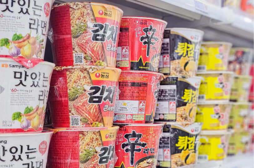 A close-up, shallow-depth-of-field shot of stacked red and yellow Nongshim and Samyang brand instant noodle cups on a store shelf.