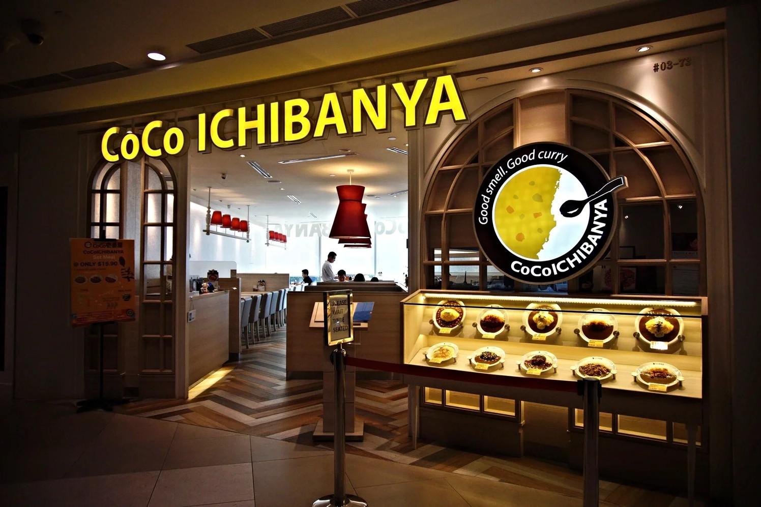  The image shows the brightly lit storefront of a CoCo Ichibanya restaurant, featuring its signature yellow signage and a circular logo that reads "Good smell. Good curry." A large display case on the right showcases several plastic food models of curry dishes, while the interior reveals a clean, modern dining area with wooden flooring and red pendant lights.