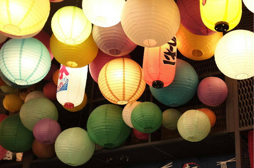A dense and vibrant cluster of colorful Japanese paper lanterns in various sizes and shades of yellow, blue, green, and pink hanging from a ceiling.