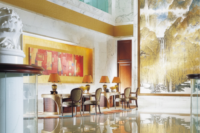 An opulent hotel lobby with marble walls, featuring gold-toned art pieces, a large waterfall mural, and elegant wooden desks with classic lamps.