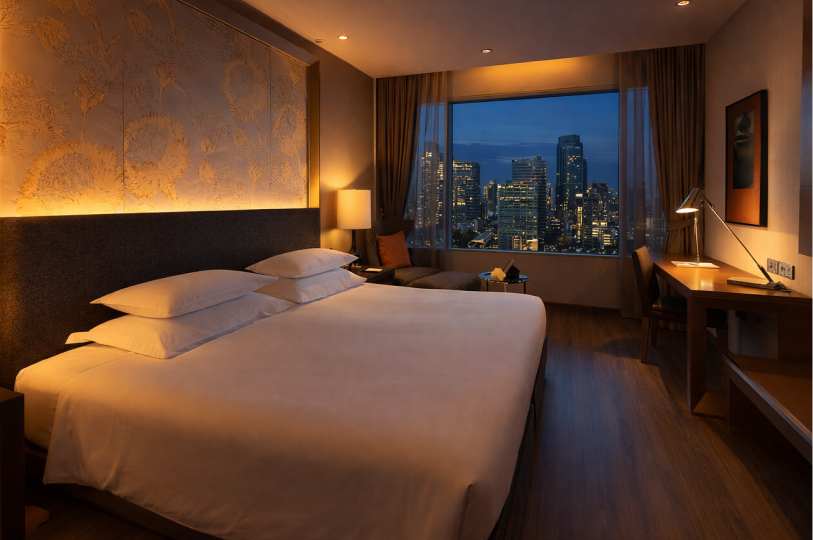 A modern hotel room with warm ambient lighting, featuring a large bed with white linens, a wooden desk, and a large window offering a night view of the Bangkok city skyline.
