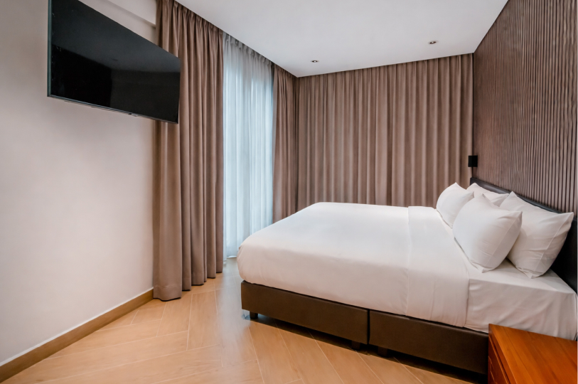 Minimalist bedroom with a king-sized bed, white linens, brown blackout curtains, and a wall-mounted TV.