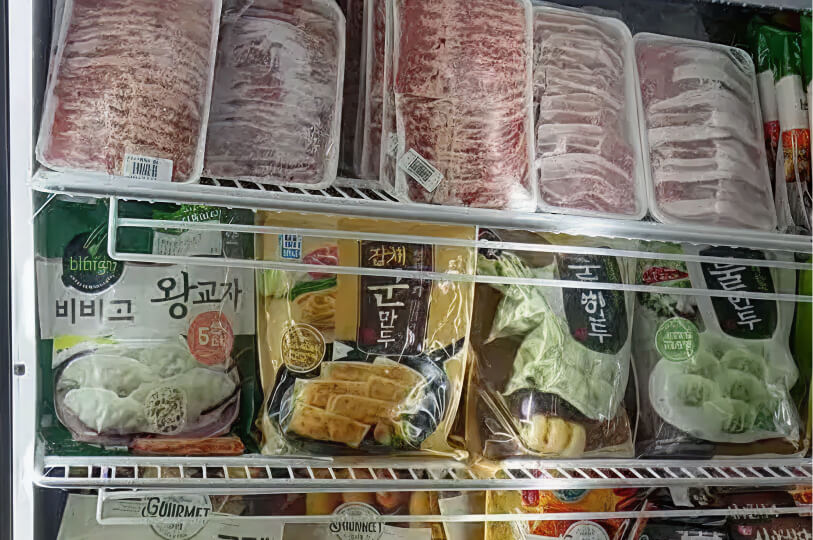 An open freezer shelf containing stacked packages of thinly sliced frozen meat and various bags of frozen Korean dumplings (mandu).
