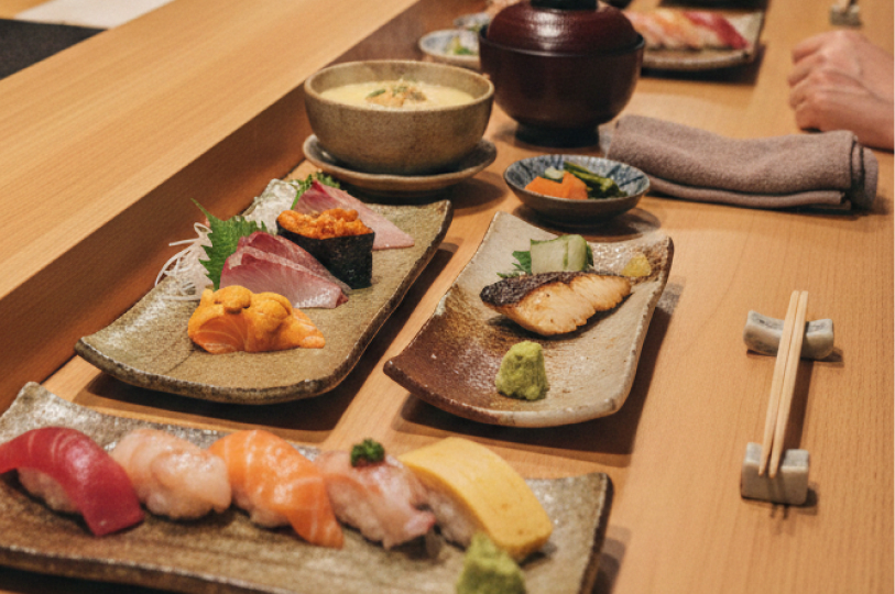 An assortment of omakase dishes on a wooden counter, including various nigiri sushi, fresh sashimi with uni, grilled fish, and a bowl of chawanmushi.