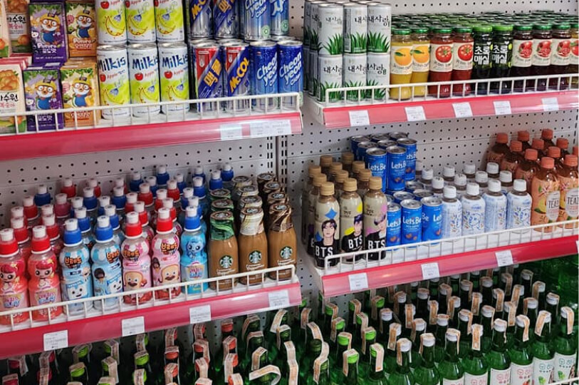 A bright display of Korean beverages on white wire shelves. The selection includes canned sodas like Milkis and Chilsung Cider, bottled juices, BTS-themed coffee drinks, and rows of green glass soju bottles on the bottom shelf.