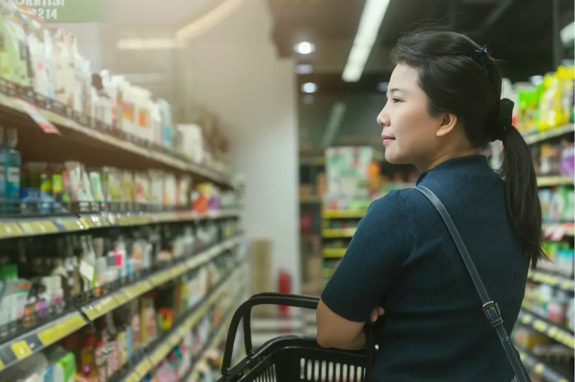A woman with her hair in a ponytail holding a shopping basket while looking at products on well-stocked grocery store shelves.