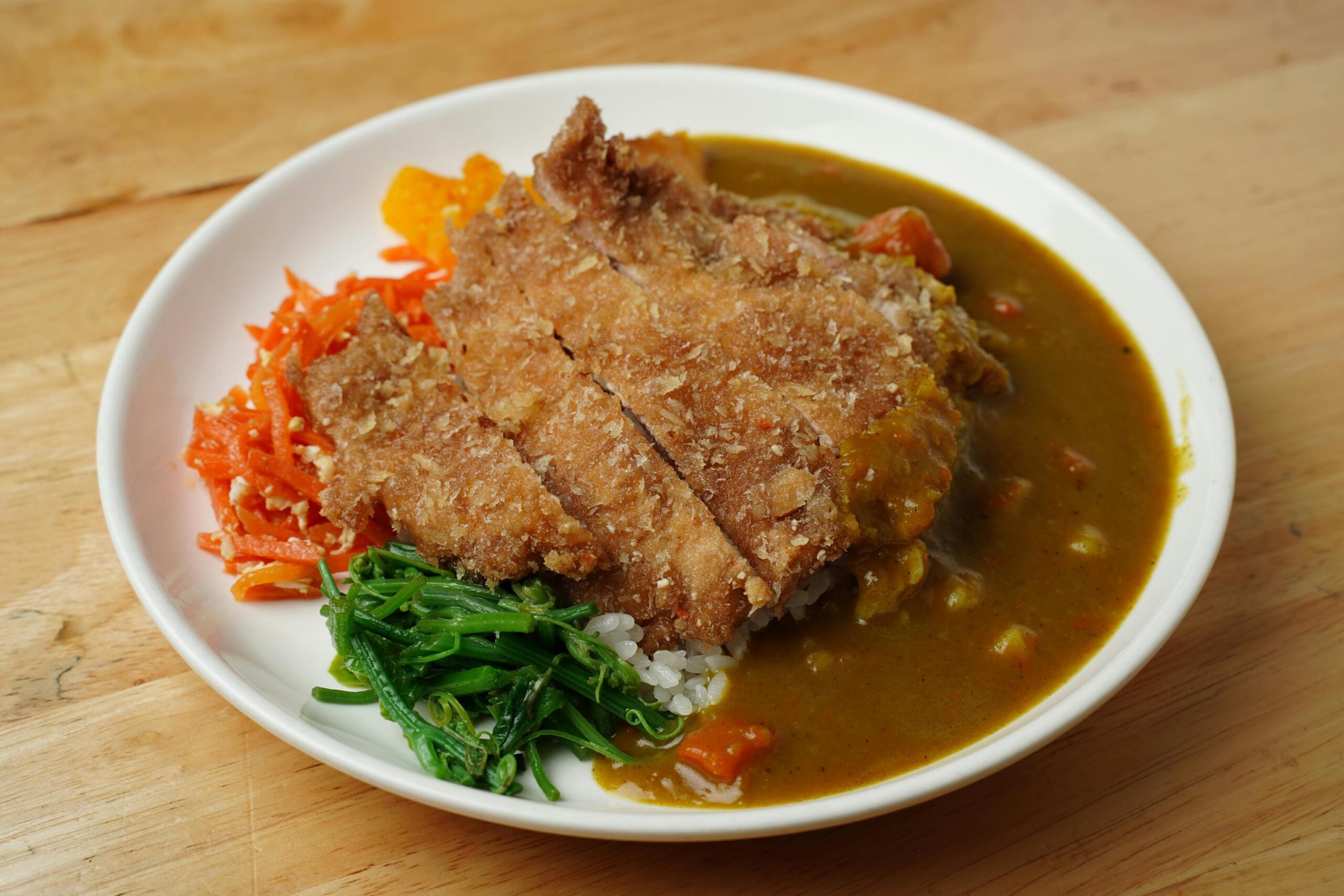 This image displays a white plate filled with a sliced breaded cutlet served over rice and a thick brown curry sauce. It is accompanied by vibrant side portions of shredded orange carrots and sautéed green leafy vegetables, all resting on a light wooden table.