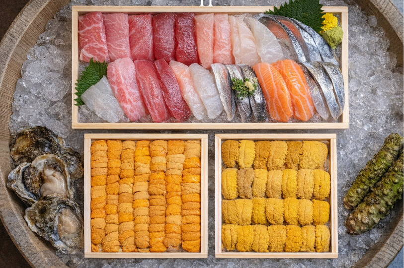 An overhead view of premium seafood arranged on ice, including wooden boxes of uni (sea urchin), assorted sliced sashimi, fresh oysters, and whole wasabi roots.