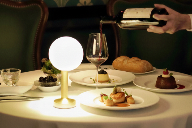 A server pouring red wine into a glass at a dimly lit, upscale dinner table featuring gourmet dishes and a glowing orb lamp.