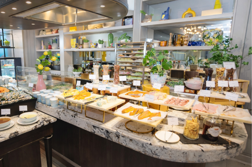 A breakfast buffet spread featuring various cheeses, cold cuts, smoked fish, and crackers on a marble countertop with shelves of decorative items in the background.