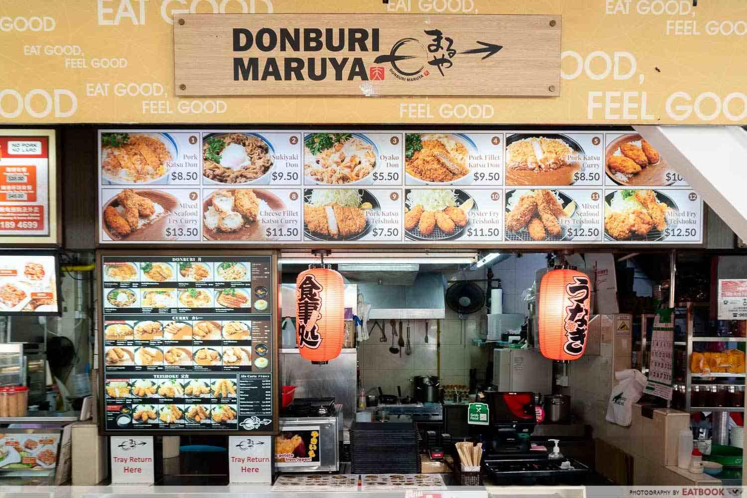 This image shows the storefront of Donburi Maruya, featuring a large menu board with photos and prices of various Japanese dishes like Pork Katsu Don and Sukiyaki Don. The stall is decorated with traditional red Japanese lanterns and includes clear signage for a tray return area and a "No Pork, No Lard" notice.