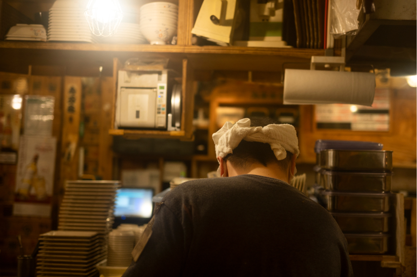 A view from behind a chef wearing a white head towel (hachimaki), working in a busy, cluttered Japanese kitchen filled with stacked bowls, plates, and cooking supplies.