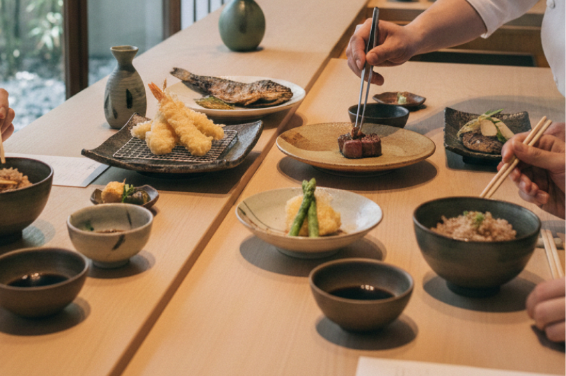 Gemini said An overhead view of a wooden dining counter featuring several Japanese dishes, including tempura prawns, grilled fish, wagyu beef, and rice bowls, with a chef's hand using tweezers to garnish a plate.