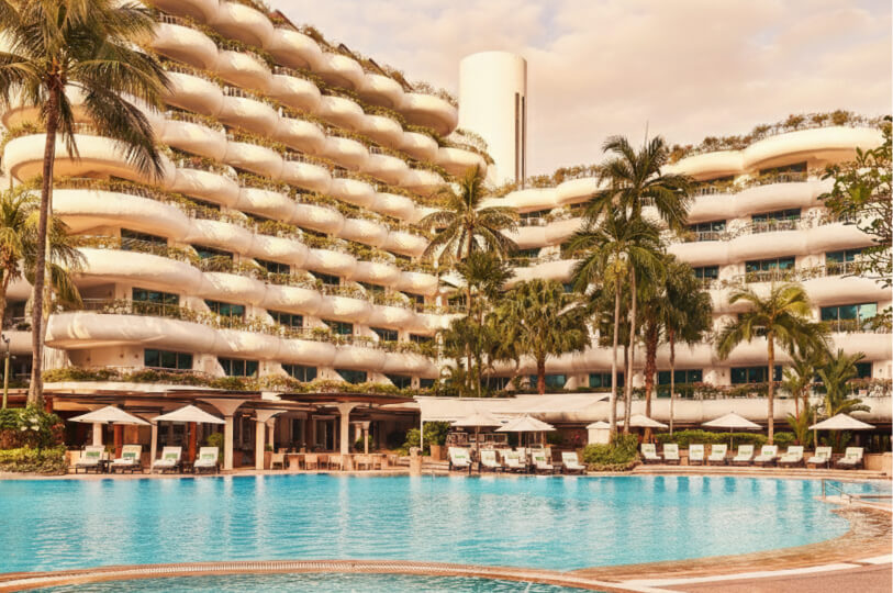 A luxury hotel resort featuring a large swimming pool and a building with distinctive rounded white balconies, framed by palm trees at sunset.