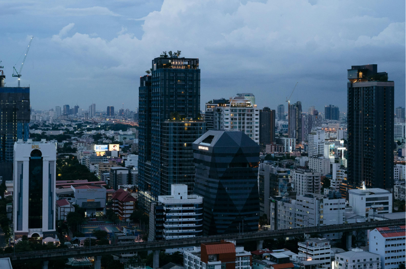 An evening aerial view of the Bangkok city skyline featuring a mix of modern skyscrapers and mid-rise buildings under a cloudy twilight sky, with a Skytrain track running through the foreground.
