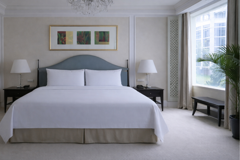 An elegant, neutral-toned hotel bedroom featuring a large bed with a soft blue headboard, flanked by dark wood nightstands and white lamps.