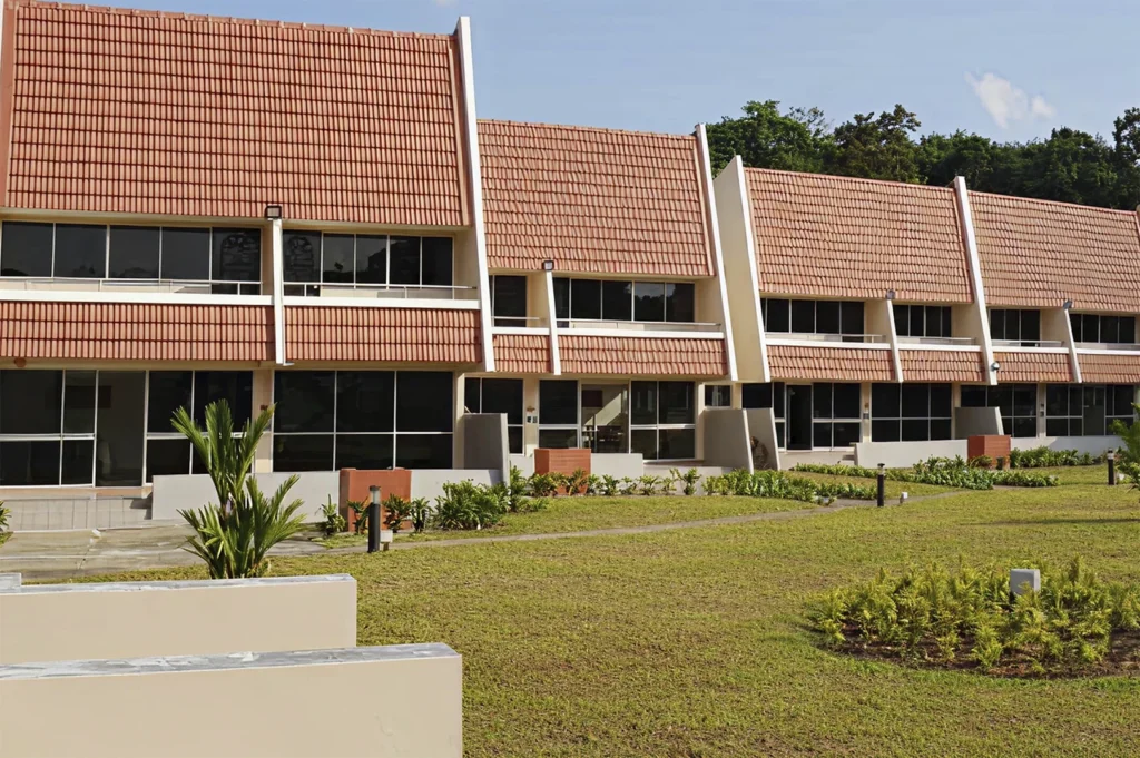 A row of two-story vacation chalets in Singapore featuring distinct, steep orange-tiled A-frame roofs, white exterior walls, and large floor-to-ceiling windows on both levels, set against a backdrop of green trees under a clear blue sky.