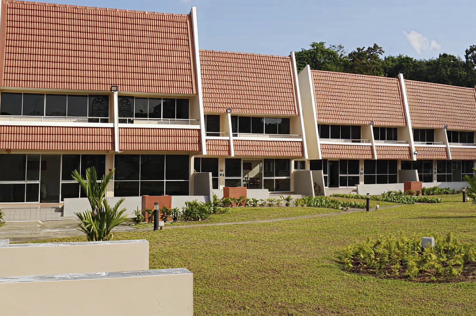 A row of two-story vacation chalets in Singapore featuring distinct, steep orange-tiled A-frame roofs, white exterior walls, and large floor-to-ceiling windows on both levels, set against a backdrop of green trees under a clear blue sky.