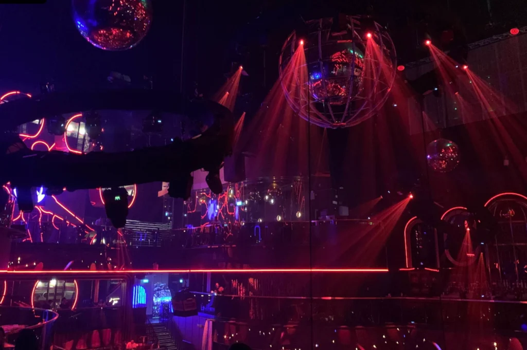 A wide-angle, low-light shot from a high vantage point inside a vibrant nightclub. Large disco balls hang from the ceiling, illuminated by intense red stage lighting that creates beams cutting through the haze. A circular structure surrounds the dance floor, and the multi-level architecture of the club is visible, drenched in deep red and purple tones.