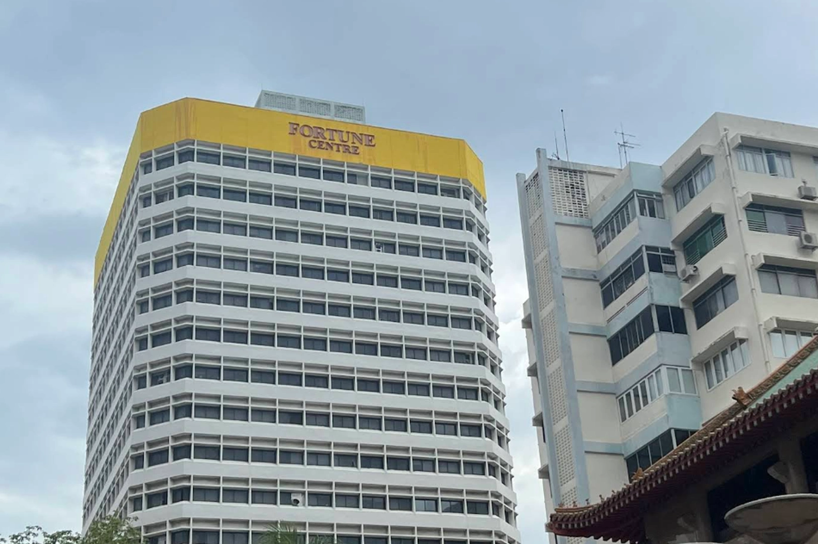 Exterior view of Fortune Centre, a tall, multi-story office building with white walls, horizontal rows of windows, and a prominent bright yellow upper section bearing the name "FORTUNE CENTRE" in bold red letters against a light blue sky.