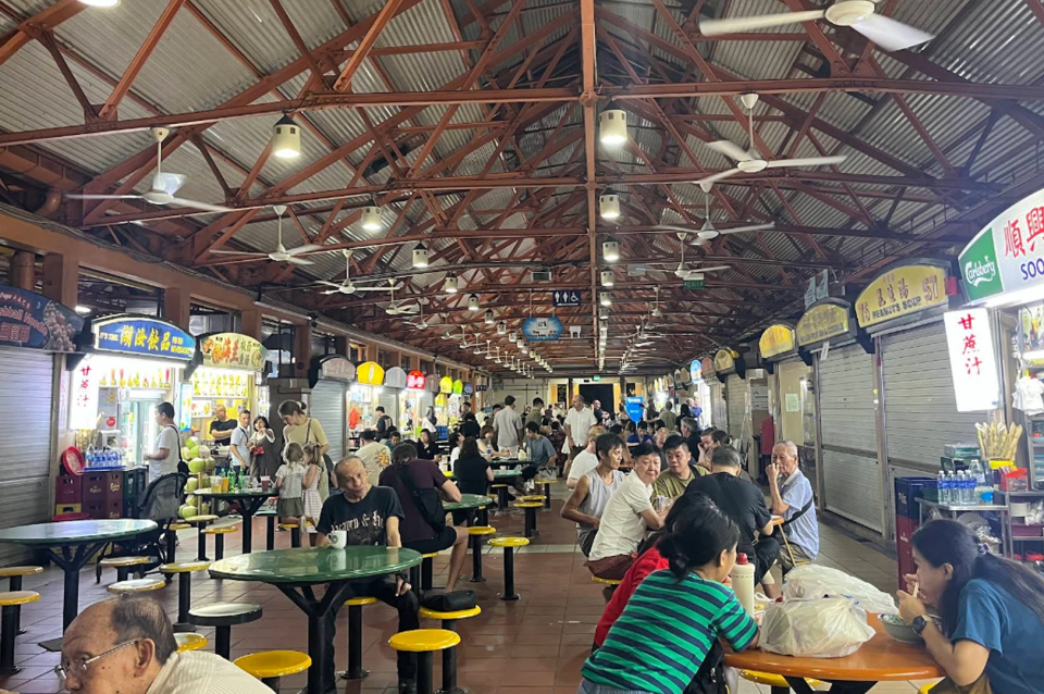 Bustling outdoor hawker center in Singapore, characterized by its industrial, high-pitched red metal truss ceiling and numerous white ceiling fans. Rows of green and orange circular tables with yellow stools are occupied by a diverse group of diners, ranging from young families to elderly individuals, creating a lively community atmosphere. The background is lined with various food stalls featuring colorful signage and bright fluorescent lighting, while the foreground shows a man in a black t-shirt seated at a green table, looking directly toward the camera. Natural light filters in from the open sides of the structure, highlighting the functional and vibrant character of this local culinary hub.
