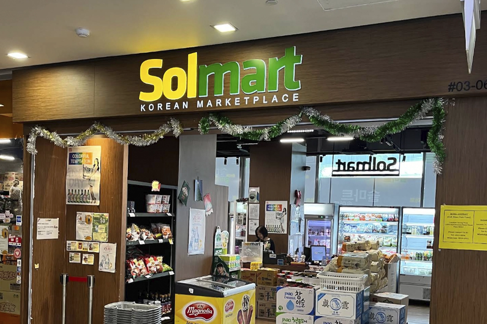 The storefront of Solmart, a Korean marketplace in Singapore. The sign features the name "Solmart" in large yellow and green letters with "KOREAN MARKETPLACE" underneath in smaller font. The entrance is decorated with green tinsel. Inside, shelves are visible with various packaged goods, a small refrigerated display unit in the foreground with "Magnolia" branding, and a cashier counter with a staff member visible in the background.