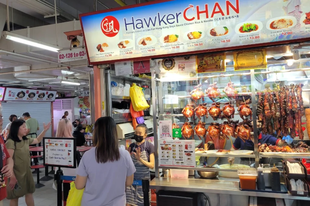 A bustling, eye-level shot of the world-famous Hawker Chan stall in a Singaporean hawker center. The bright red and white signage prominently displays the brand name and a menu featuring Soya Sauce Chicken Rice and Noodles with prices ranging from $3.50 to $4.50. A row of whole, glossy, roasted soya sauce chickens hangs behind a glass display case alongside strips of char siew. A female staff member wearing a black mask and a striped apron stands at the counter, gesturing while interacting with a customer whose back is to the camera. To the left, several people are seen in the background near red dining tables, under the industrial fluorescent lighting of the food center. The scene is candid, capturing the authentic, busy atmosphere of a popular food destination.