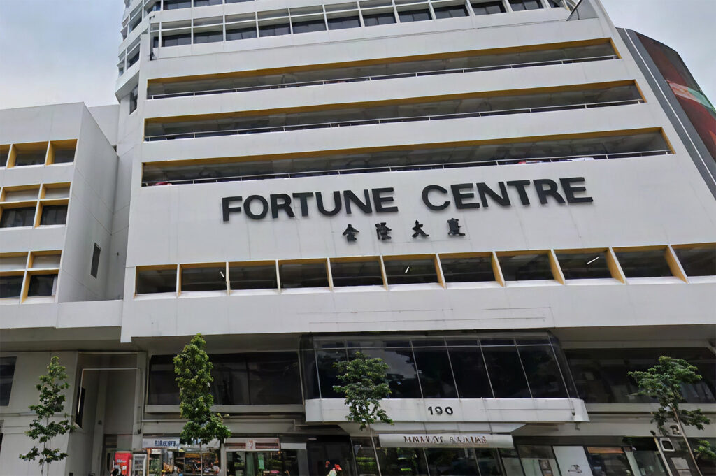 A low-angle shot of the Fortune Centre building facade in Singapore. The white concrete building features the name in large black block letters with Chinese characters below it. Small green trees line the sidewalk at the base.