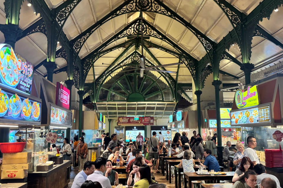 Bustling atmosphere of the Lau Pa Sat festival market in Singapore. The historic structure is defined by its iconic Victorian-era cast-iron architecture, featuring intricate dark green filigree arches and soaring cream-colored vaulted ceilings. A large industrial ceiling fan hangs from the central apex. The space is filled with a diverse crowd of people seated at long wooden communal tables, engaged in conversation and dining. Surrounding the central seating area are various brightly lit hawker stalls with colorful signage, including "Lau Pa Sat Laksa" and "Indian Cuisine." The lighting is a mix of warm glow from the stalls and soft ambient light filtering through the high glass windows in the background. The floor is made of rustic wooden planks, and the overall scene feels lively, authentic, and culturally vibrant.