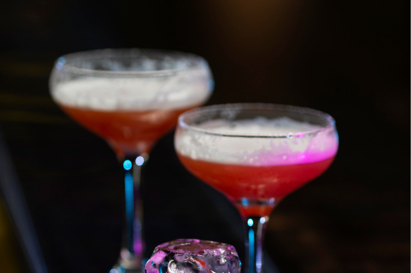 Two frosted coupe glasses filled with a pink, foamy cocktail, set against a dark background with blue and purple accent lighting.
