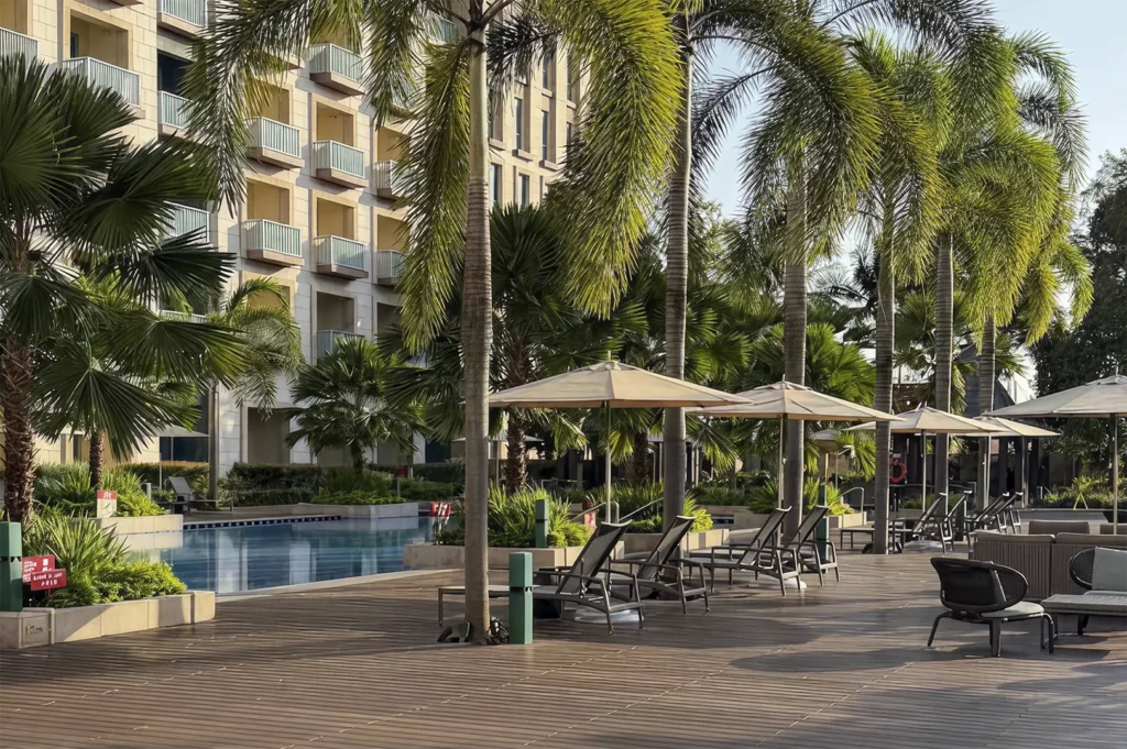 A high-angle, first-person perspective shot of a sun-drenched hotel pool deck. In the foreground, a dark wood-planked terrace is patterned with long, sharp shadows from nearby palm trees. Several grey lounge chairs sit under beige patio umbrellas. To the left, the edge of a clear blue swimming pool is visible, bordered by lush green tropical plants. In the background, the modern multi-story facade of Hotel Ora features numerous small, light-colored balconies under a bright, clear sky.