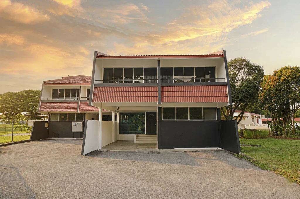 A wide shot of a modern, two-story beach chalet at dusk. The building features a distinctive sloped red roof and a dark grey lower exterior. A paved driveway sits in front, and a vibrant sunset with orange and yellow clouds fills the sky.