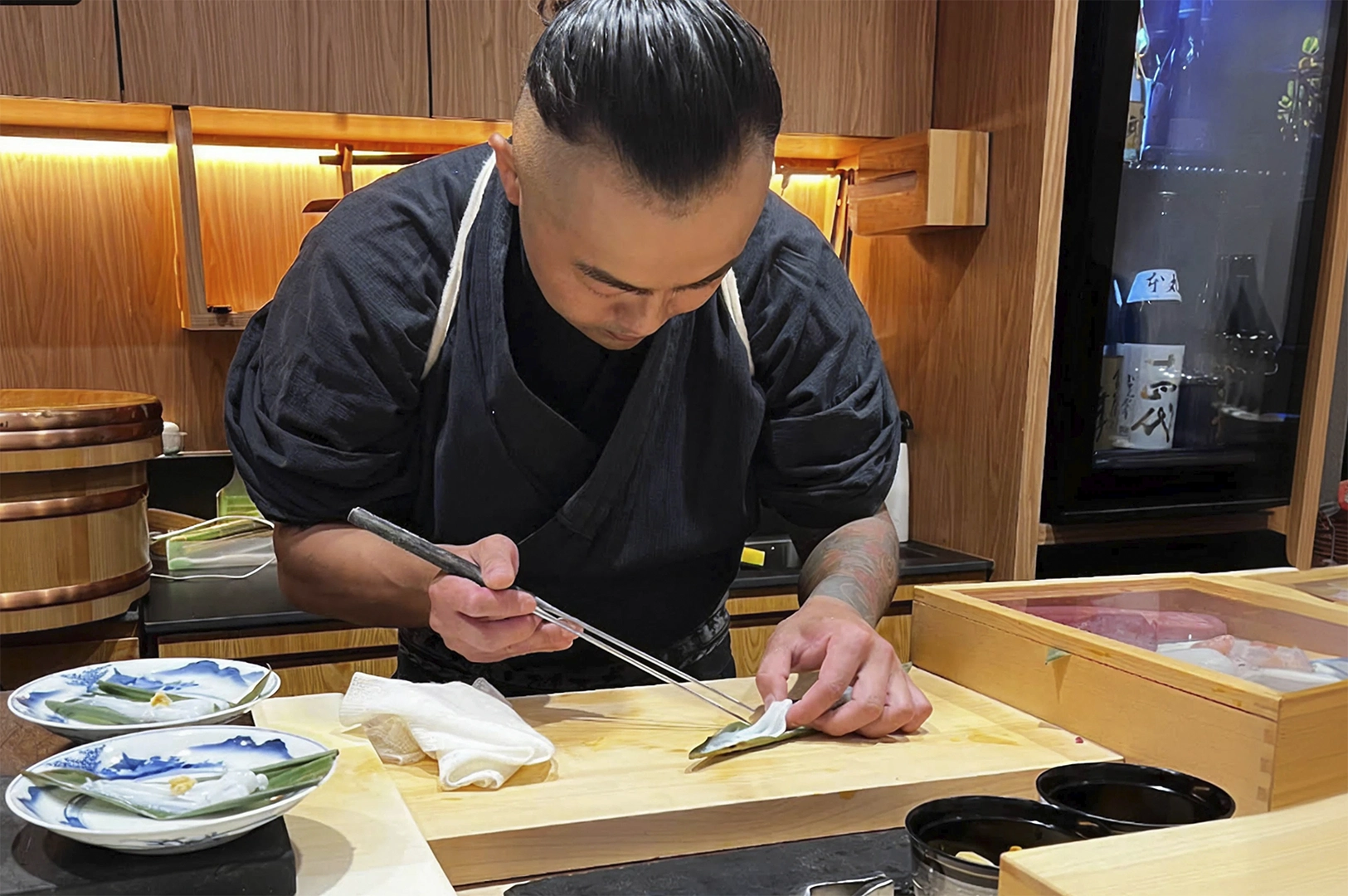 A candid, eye-level shot of a sushi chef with a faded undercut hairstyle and a tattooed arm, meticulously preparing a dish at a wooden counter. He uses long metal cooking chopsticks to carefully arrange a translucent piece of seafood on a green leaf. To his left are small blue-and-white patterned plates, and to his right is a wooden box containing various cuts of fresh fish. The background features warm wood cabinetry and a professional kitchen setting.