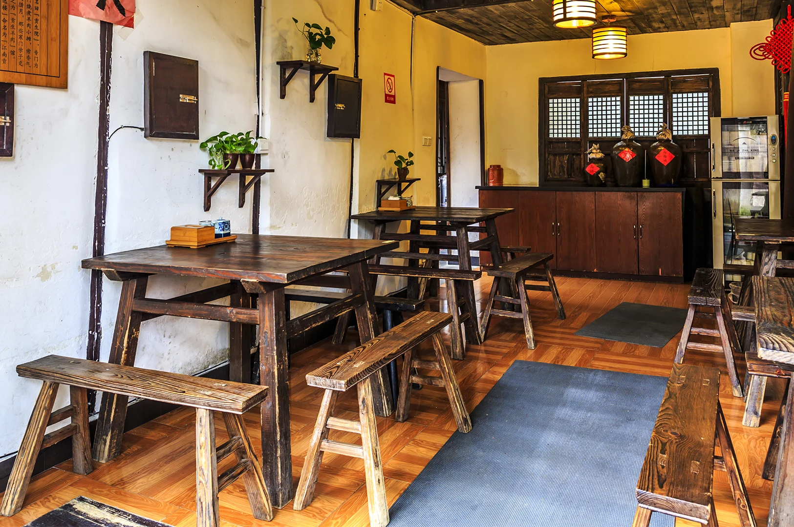 The interior of a rustic, traditional-style cafe featuring warm lighting and dark wooden furniture. In the foreground, a large wooden table with matching benches is set with a small, wooden condiment box. In the background, similar tables and benches are arranged, leading toward a wooden counter area decorated with three large, dark ceramic jugs with red labels. A vintage-style wooden window frame with a grid pattern is mounted on the back wall, and several small potted green plants are placed on wall-mounted shelves, creating a cozy, lived-in atmosphere