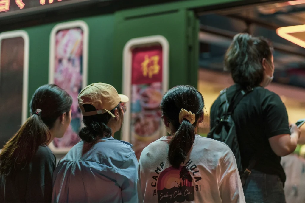 A medium shot taken from behind shows four people standing in a line, waiting to enter a restaurant. In the foreground, three individuals are captured from the back: a person with long dark hair tied in a low ponytail, a person wearing a beige baseball cap and light blue shirt, and a person with a dark ponytail wearing a white long-sleeved shirt with a vintage-style graphic of a rainbow and palm trees. To the far right, a fourth person stands partially out of frame, looking at a smartphone. They are queuing in front of a storefront with a dark green facade featuring lit, vertical menu panels or posters featuring food images and Chinese characters. The lighting is warm and somewhat dim, suggesting an evening setting.